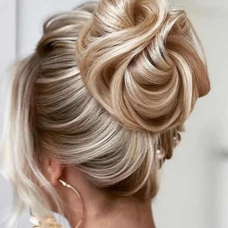 Eine Frau mit blondem Haar in einem Dutt mit losen Strähnen, die ihr Gesicht umrahmen, trägt Perlenohrringe und Creolen.