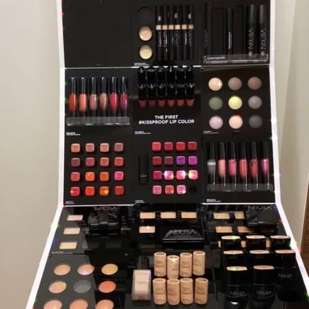 Eine Kosmetikanzeige präsentiert verschiedene Make-up-Produkte wie Lippenstifte, Lidschatten und Foundation in verschiedenen Farben. Der Markenname 'NOUVA' ist prominent sichtbar.