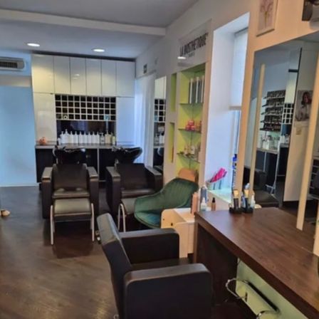 Ein Innenraum eines Salons mit Holzboden, weißen Wänden und mehreren schwarzen Stühlen. Vor einem Spiegel steht ein Holzschreibtisch. Regale mit Flaschen und Produkten sind sichtbar.