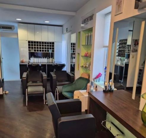 Ein Innenraum eines Salons mit Holzboden, weißen Wänden und mehreren schwarzen Stühlen. Vor einem Spiegel steht ein Holzschreibtisch. Regale mit Flaschen und Produkten sind sichtbar.