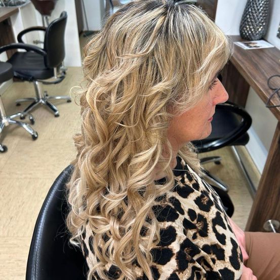 Eine Frau mit blonden Locken sitzt in einem Salonstuhl und trägt ein Leoparden-Print-Oberteil. Hinter ihr ist ein Holzschreibtisch und zwei schwarze Drehstühle zu sehen.