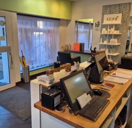 Ein Blick in das Innere eines Geschäfts mit Schreibtisch, Monitor, Tastatur und Maus. Das Geschäft hat eine Glastür, ein Regal mit Flaschen und ein Schild mit der Aufschrift KEUNE.