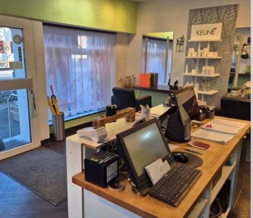 Ein Blick in das Innere eines Geschäfts mit Schreibtisch, Monitor, Tastatur und Maus. Das Geschäft hat eine Glastür, ein Regal mit Flaschen und ein Schild mit der Aufschrift KEUNE.