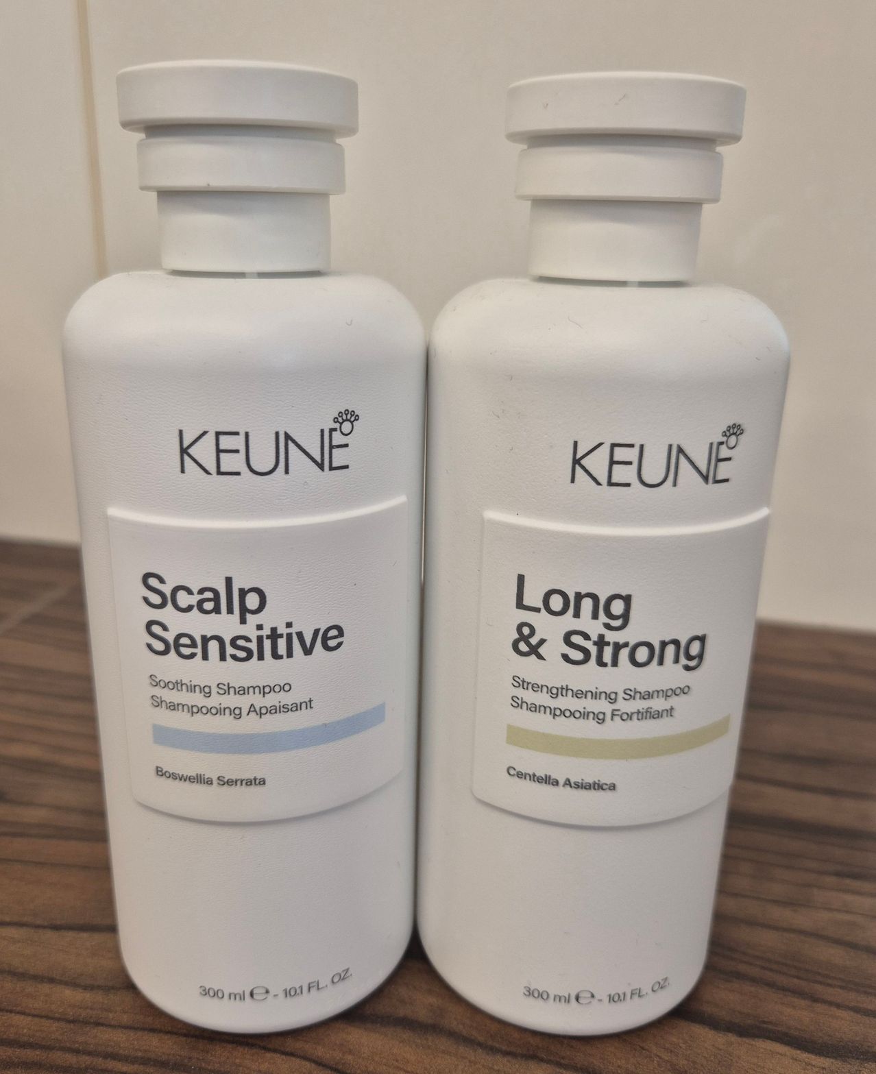 Two bottles of Keune shampoo on a wooden surface. One is labeled Scalp Sensitive and the other Long & Strong.