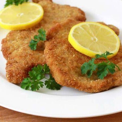 Drei Stücke paniertes Fleisch, jedes mit einer Zitronenscheibe und Petersilie garniert, auf einem weißen Teller mit einer Holzoberfläche.