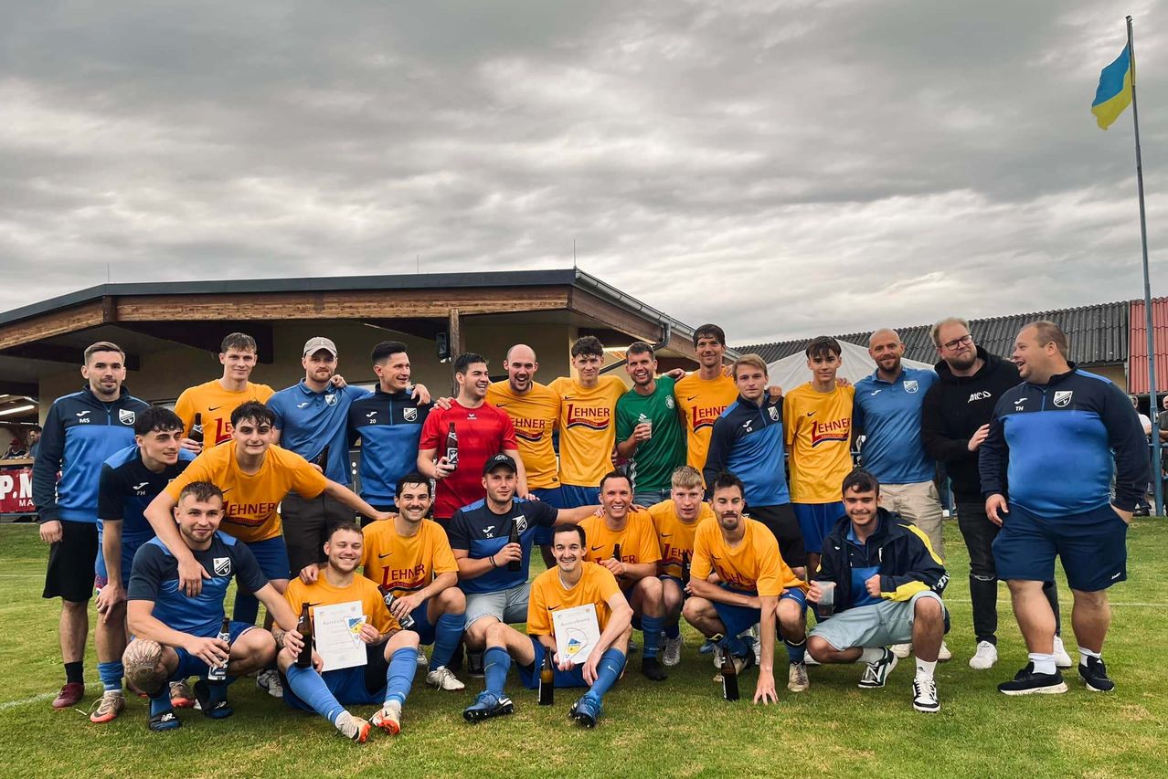 Eine Gruppe von Fußballspielern posiert für ein Foto vor einem Gebäude mit bewölktem Himmel. Einige tragen Trikots in Gelb und Blau, während andere Freizeitkleidung tragen. Einige halten Bierflaschen, andere halten Zertifikate.