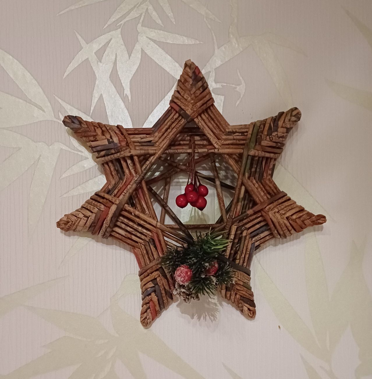 Eine sternförmige Weihnachtsdekoration aus Zweigen mit roten Beeren und Tannenzapfen hängt an einer weißen Wand mit Bambusmuster.