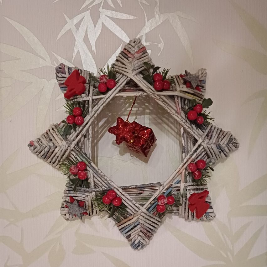 Ein sternförmiger Kranz aus geflochtenem Stroh, verziert mit roten Beeren und grünen Blättern. In der Mitte hängt eine rote Geschenkbox an einem Band. Der Hintergrund ist eine weiße Tapete mit einem Bambusmuster.