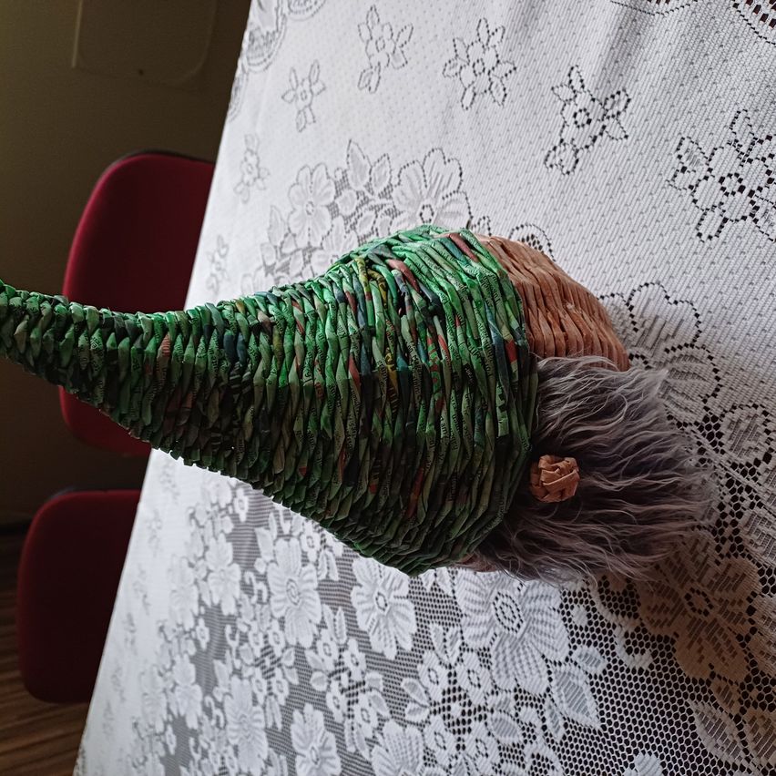 Ein handgefertigter Zwerg mit einem grünen kegelförmigen Hut sitzt auf einer Tischdecke mit Blumenmuster, in der Nähe roter Stühle, mit Licht, das von links beleuchtet wird.