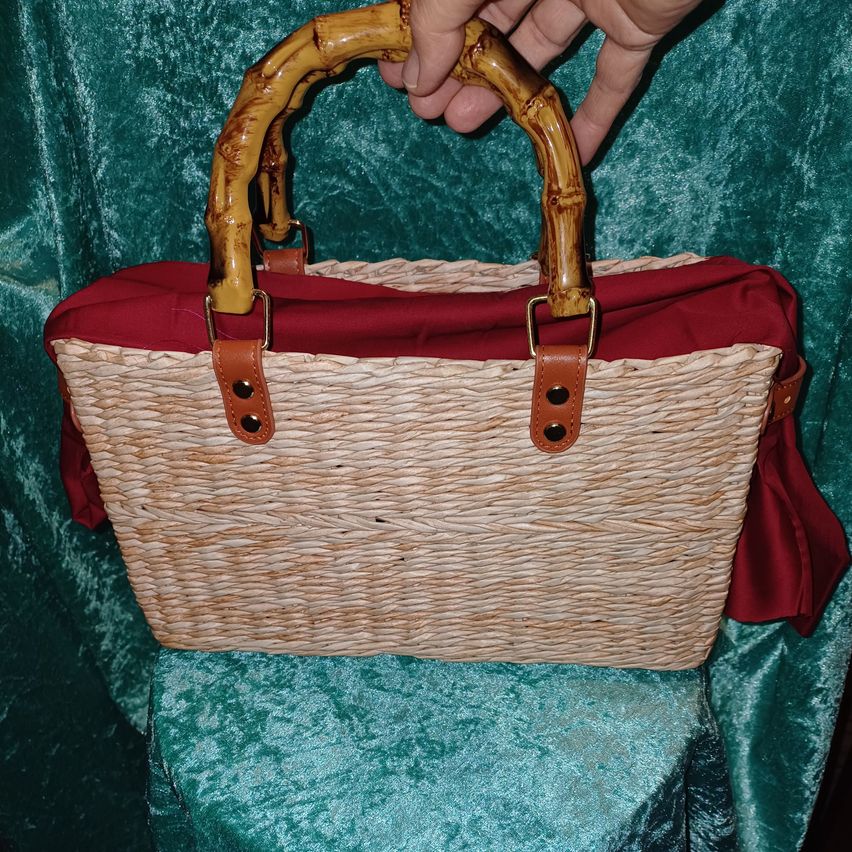 Eine Person hält eine Korbtasche mit Bamushenkeln, über die ein roter Stoff gelegt ist, auf einer blaugrünen Samtfläche.