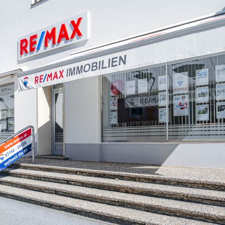 Ein RE/MAX Immobilien Ladenlokal mit Glasfenstern und einem Banner über dem Eingang. Das Gebäude hat Steinstufen, die hinaufführen.