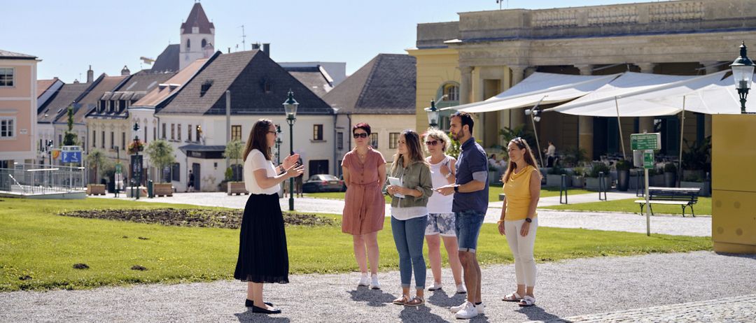 Eine Frau führt eine Gruppe von Touristen auf einer Kopfsteinpflasterstraße vor einem alten Gebäude mit verschiedenen Architekturstilen.