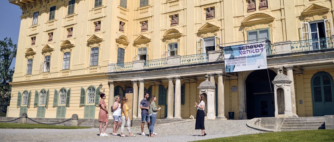 Eine Gruppe von Menschen steht vor einem großen gelben Gebäude mit klassischer Architektur. Das Gebäude hat viele Fenster und Statuen. Eine Fahne ist an der Seite aufgehängt.