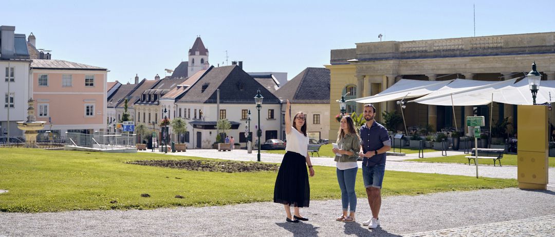Drei Personen stehen auf einer gepflasterten Straße, eine Frau zeigt nach oben. Sie stehen vor einem Gebäude mit vielen Fenstern und einem Turm. Im Hintergrund befinden sich weitere Gebäude und eine Rasenfläche.