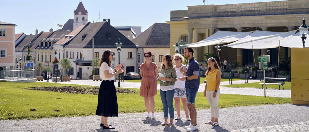 Eine Frau in einem weißen Oberteil und einer schwarzen Schürze spricht vor einer Gruppe von Menschen. Sie stehen auf einem Kopfsteinpflaster, in der Nähe eines Rasens, mit mehreren Gebäuden im Hintergrund.