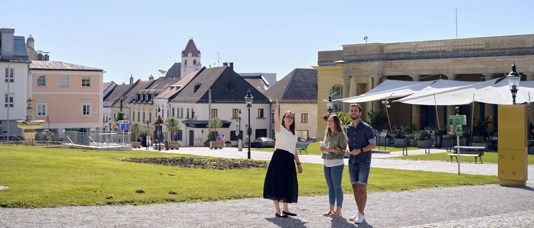 Drei Menschen stehen auf einem gepflasterten Platz vor Gebäuden, eine Frau zeigt nach oben. Hinter ihnen ein grüner Rasen und einige Bäume. Rechts ein Schirm. Der Himmel ist klar.
