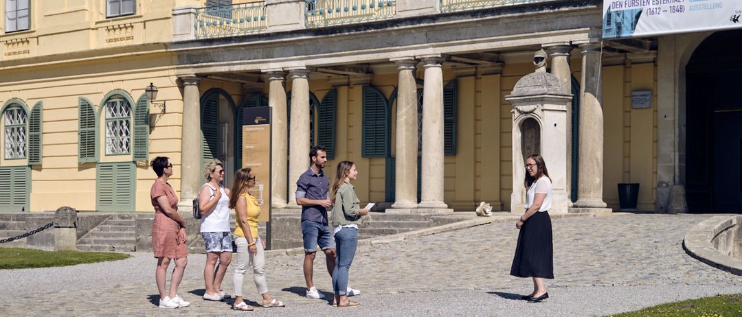 Eine Gruppe von Menschen steht vor einem historischen Gebäude mit einem Schild über die sieben Fürsten. Das Gebäude hat eine klassische Architektur mit Säulen und Fenstern.