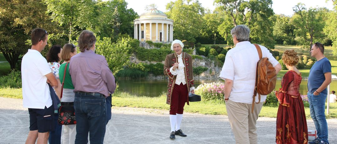 Ein älterer Mann in historischer Kleidung hält eine Präsentation in einem Park. Mehrere Menschen hören zu. Ein klassisches Gebäude ist im Hintergrund zu sehen.