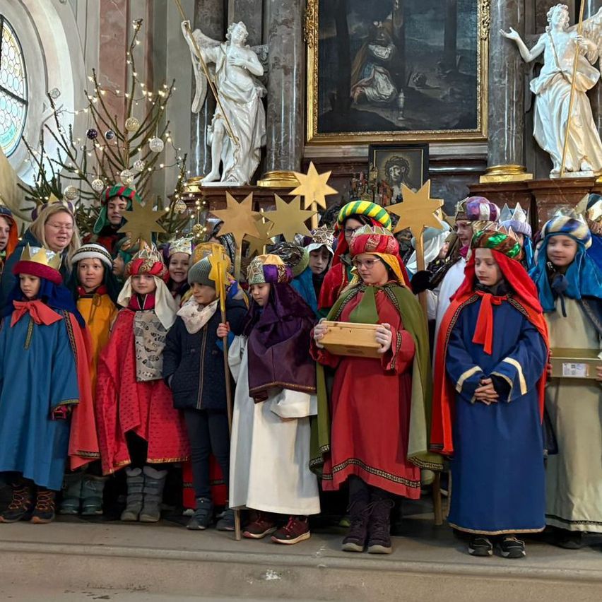 Eine Gruppe von Kindern in traditionellen Kostümen und Mützen steht in einer Kirche und hält Sterne. Sie posieren für ein Foto.