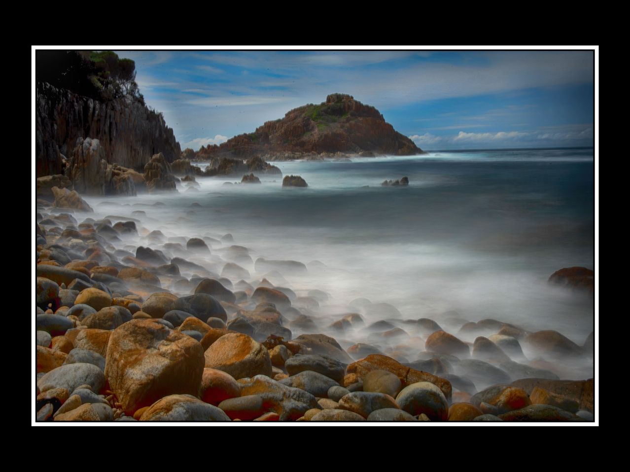 Eine ruhige Küstenszene mit einem felsigen Ufer, verstreuten Steinen und einer kleinen Insel in der Ferne unter einem teilweise bewölkten Himmel.