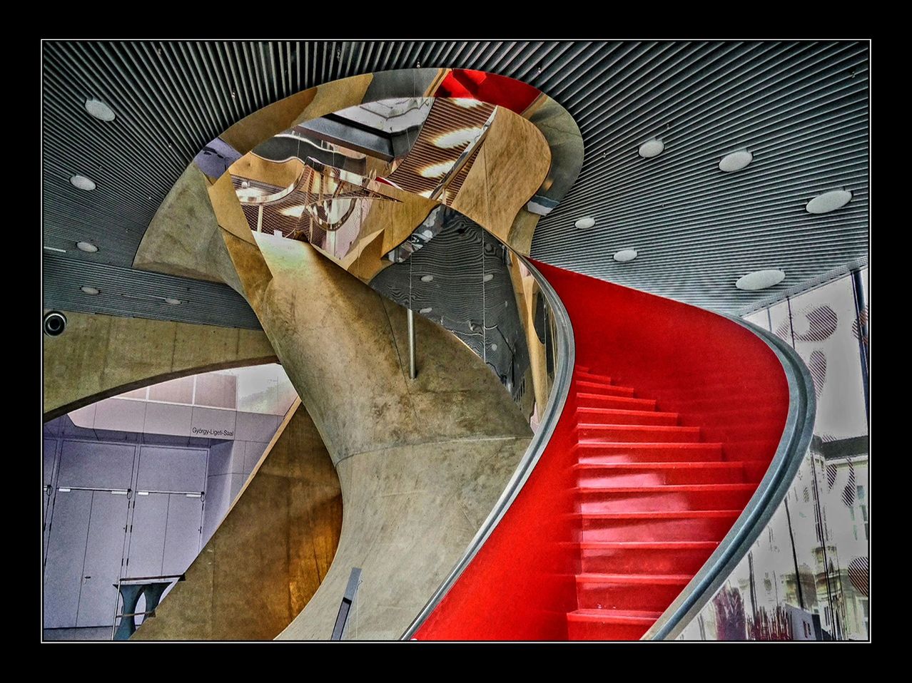 A modern spiral staircase with red steps, metallic railing, and reflective surfaces. The structure has a geometric design, with a gray ceiling and a reflection of the upper level.