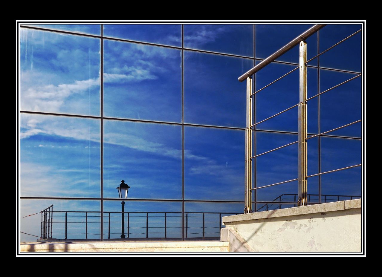 Ein modernes Gebäude mit Glaswänden, das einen blauen Himmel und Wolken reflektiert. Eine schwarze Straßenlampe steht auf einem Balkon mit einem Geländer.