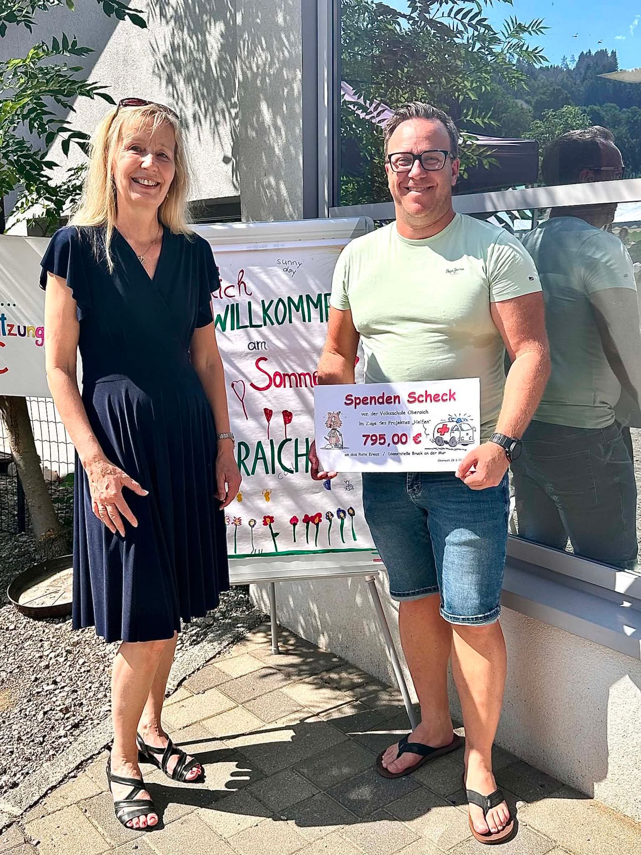 Eine Frau und ein Mann stehen neben einem Banner mit der Aufschrift 'Willkommen am Reich' und halten einen Scheck über 795,00 Euro. Beide tragen Freizeitkleidung und lächeln für die Kamera. Hinter ihnen spiegelt eine Glasscheibe einen Baum und eine weitere Person.