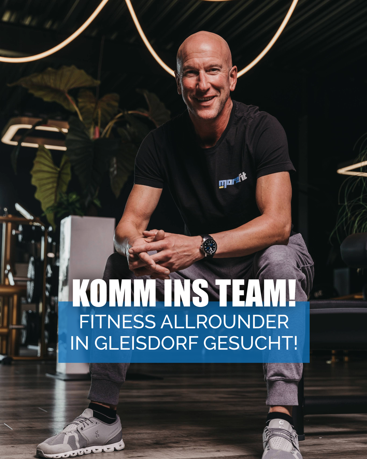 Ein Mann im Fitnessstudio trägt ein schwarzes T-Shirt und graue Hose. Er sitzt lächelnd. Ein Text-Overlay lautet 'Komm ins Team! Fitness Allrounder in Gleisdorf gesucht!'.