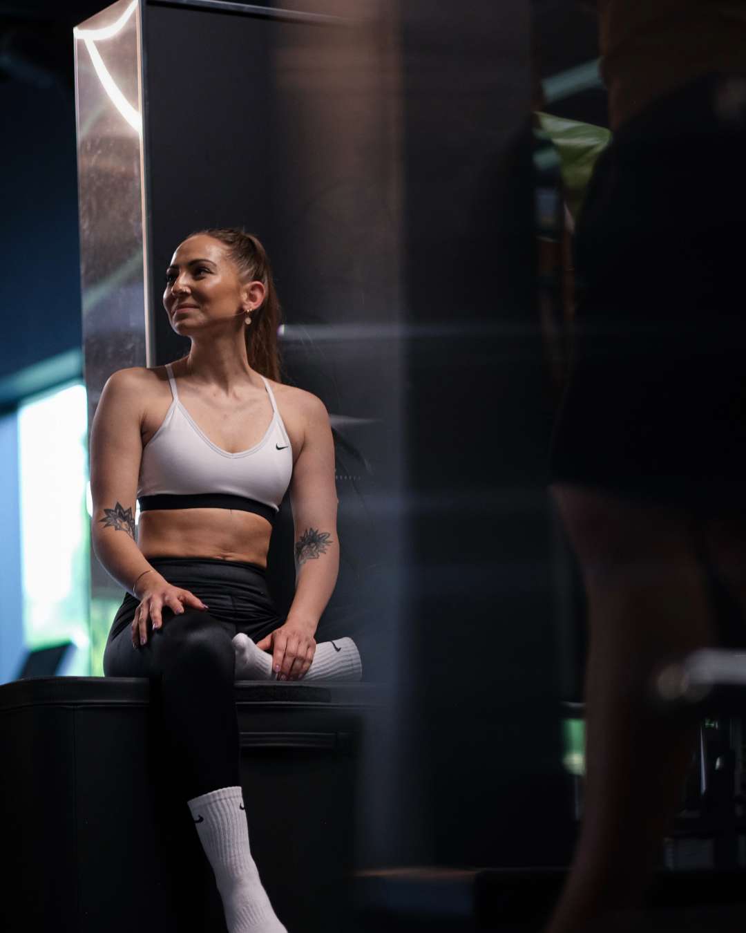 Eine Frau in Trainingskleidung sitzt auf einer Bank im Fitnessstudio. Sie hat Tattoos an den Armen und lächelt in die Kamera.