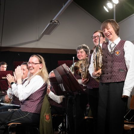 Mitglieder einer Band klatschen und lächeln, während sie auf der Bühne stehen. Die Frau in der Mitte hält ein Blechblasinstrument.