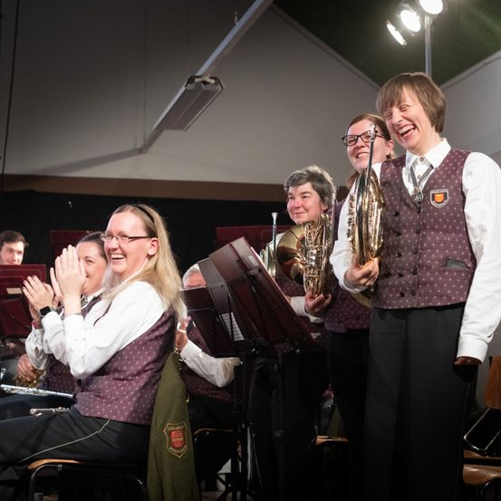 Mitglieder einer Band klatschen und lächeln, während sie auf der Bühne stehen. Die Frau in der Mitte hält ein Blechblasinstrument.