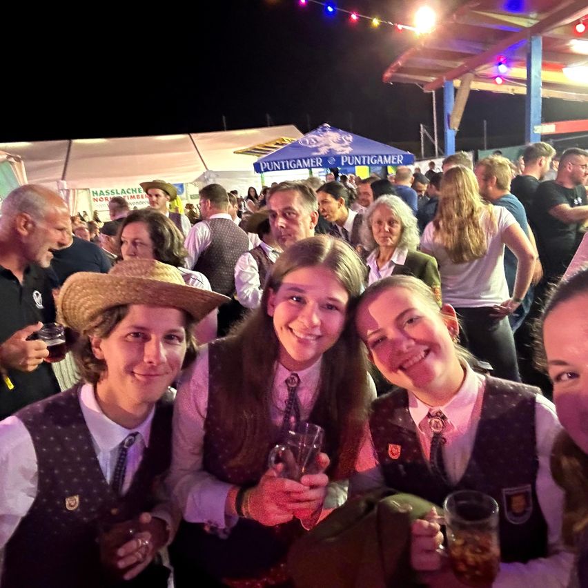 Eine Gruppe von Menschen posiert für ein Foto auf einem Festival bei Nacht. Eine Frau mit Strohhut und ein Mädchen mit Abzeichen lächeln. Sie halten Getränke.