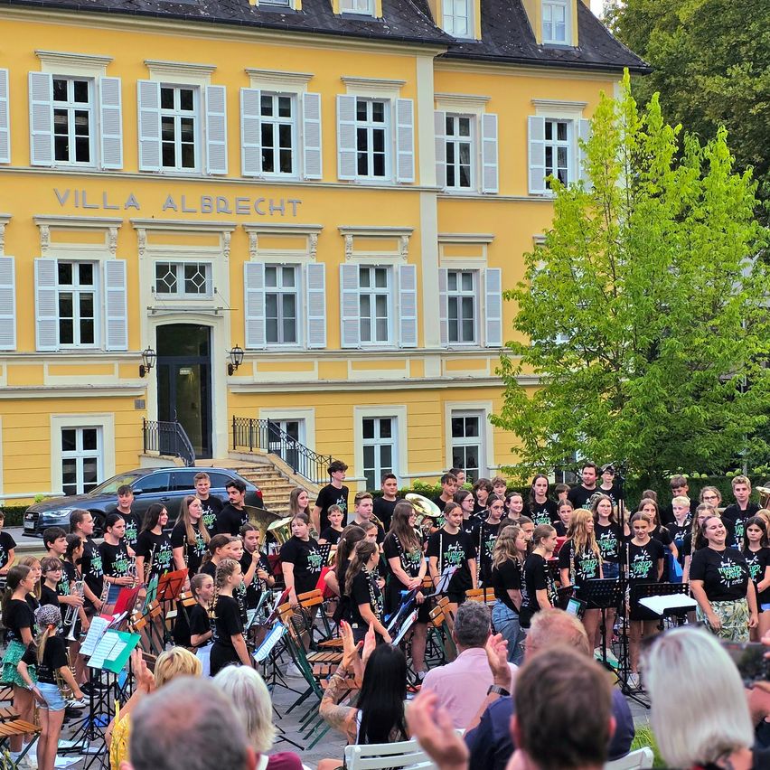 Ein Blasorchester spielt vor dem gelben Gebäude der Villa Albrecht. Viele Menschen sehen sich die Aufführung an.