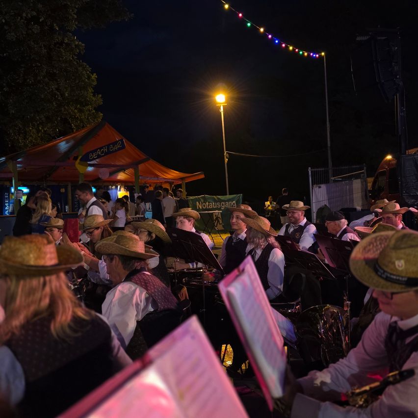 Eine Gruppe von Musikern in Strohhüten spielt bei einer nächtlichen Veranstaltung unter einem Zelt mit bunten Lichtern.