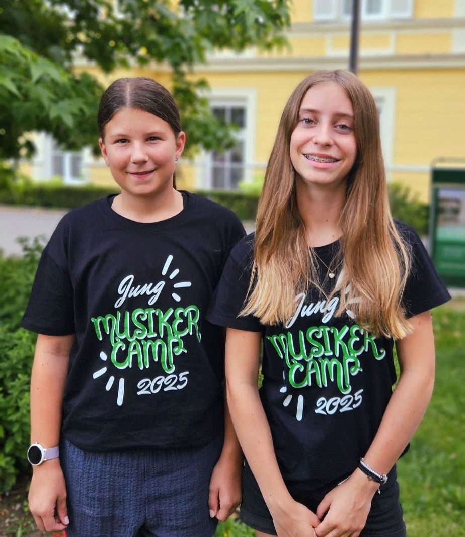 Zwei junge Mädchen tragen schwarze T-Shirts mit dem Text 'Jung Musiker Camp 2025' und stehen vor einem gelben Gebäude mit Pflanzen und Bäumen.