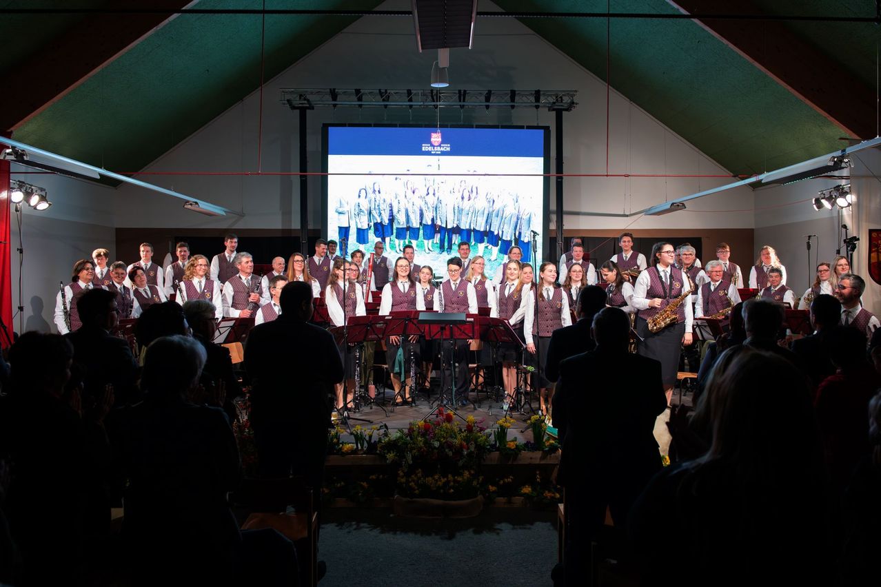 A choir and orchestra perform on stage in front of a large screen and an audience. The stage is decorated with flowers.
