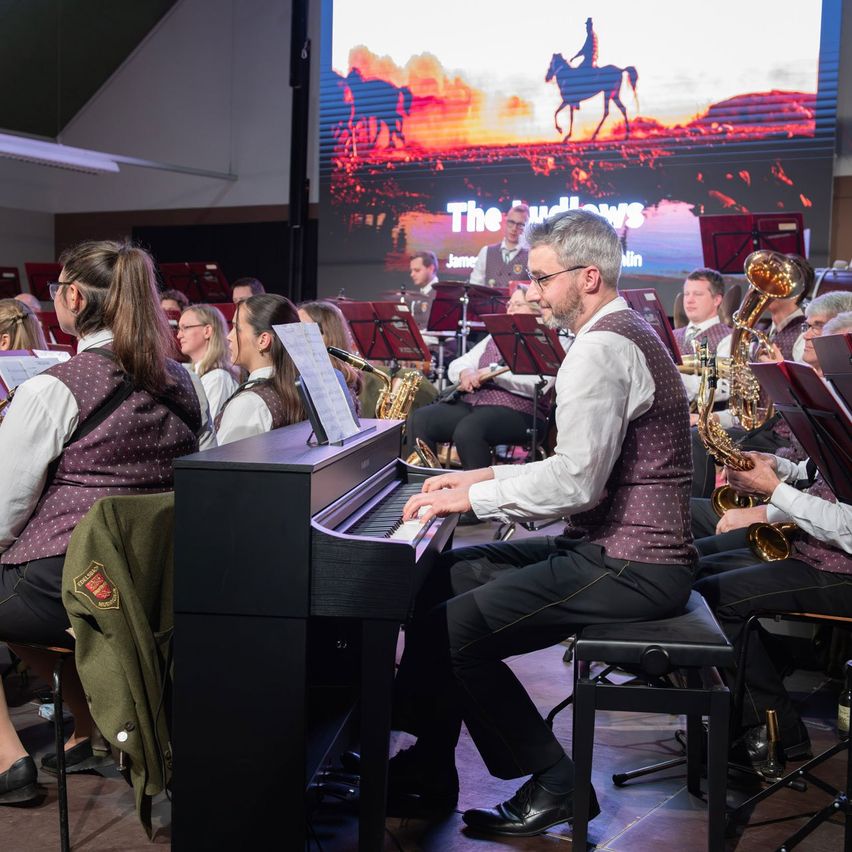 A band with a pianist, saxophone players, and other musicians performs in a hall with a large screen showing a sunset and a horse rider.