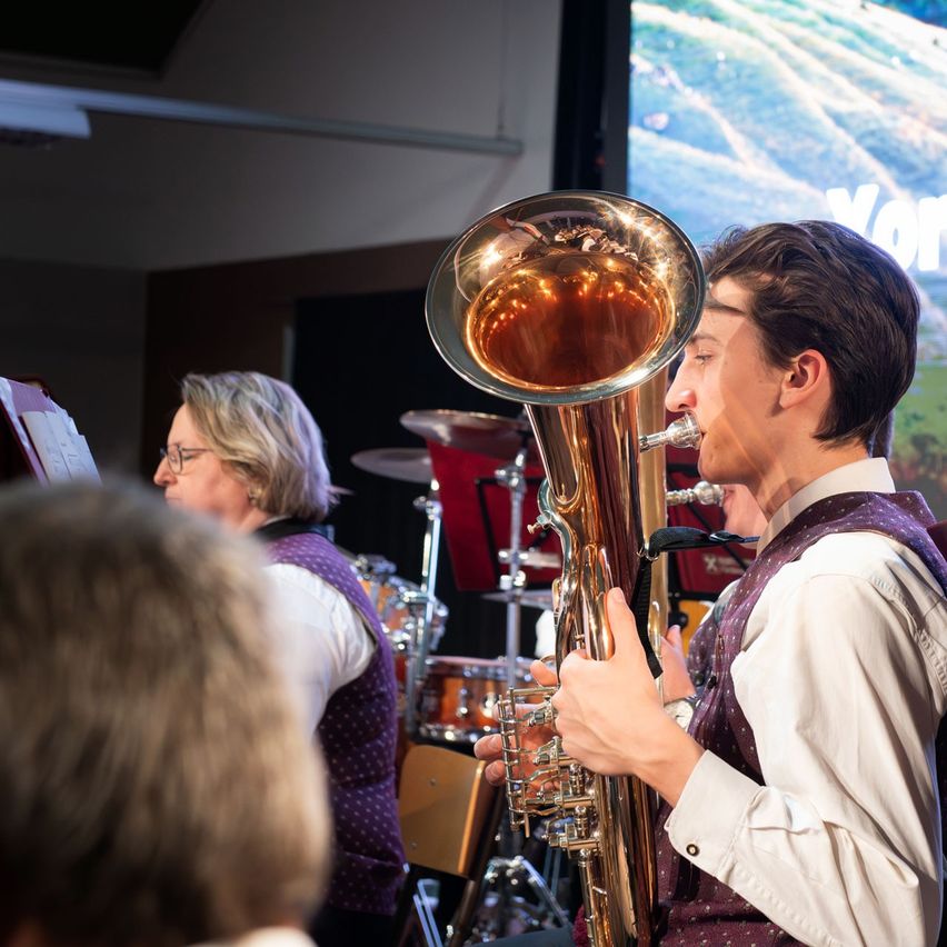 A man plays a tuba at a concert while a woman reads sheet music in the background. The audience watches the performance.