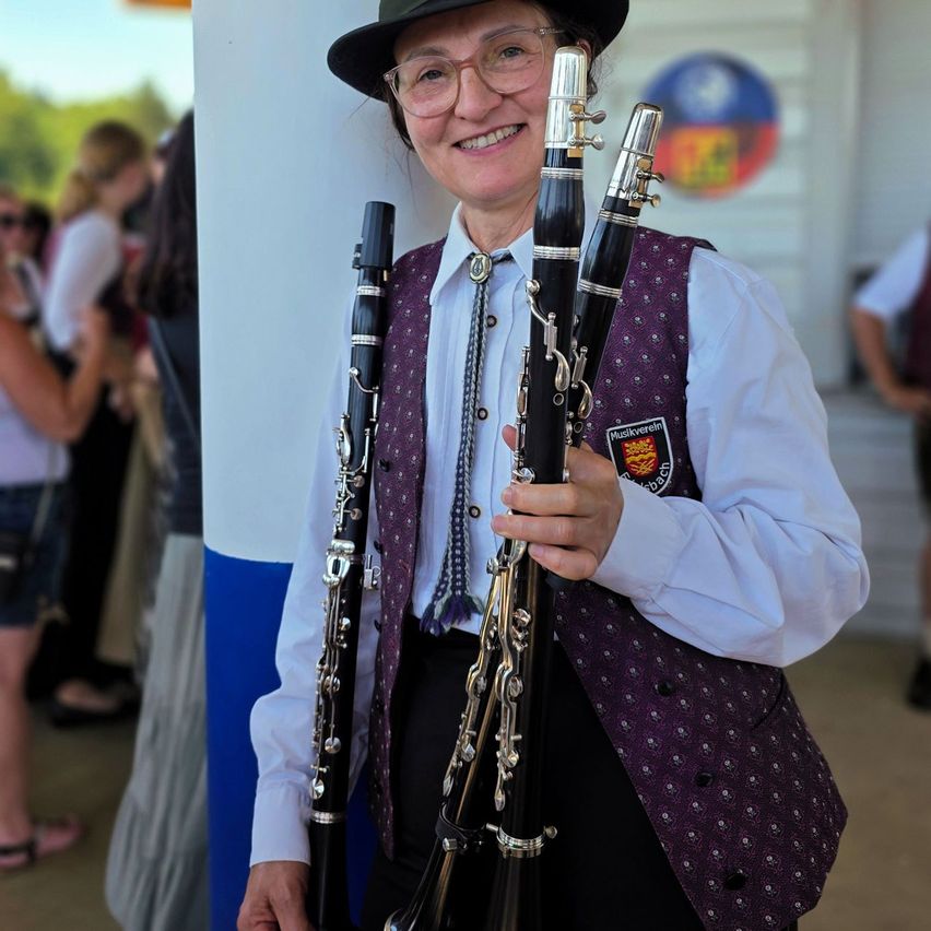 A woman in a purple vest holds two clarinets, smiling for the camera in an outdoor setting. Behind her, blurred figures and a blue-and-white striped pole are visible.