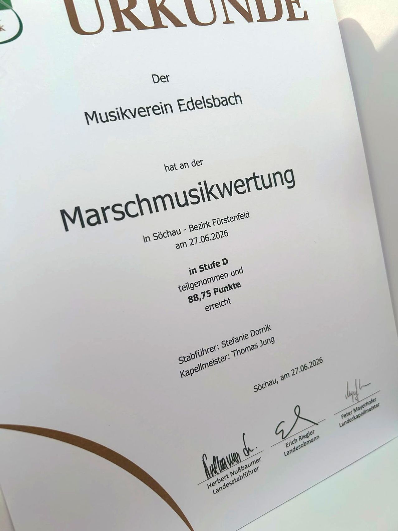 A white sheet with black text is displayed, reading 'Der Musikverein Edelsbach'. It includes details about a music evaluation event held in SÃ¶chau at the FÃ¼rstenfeld district on 27.06.2026, awarding 88.75 points.