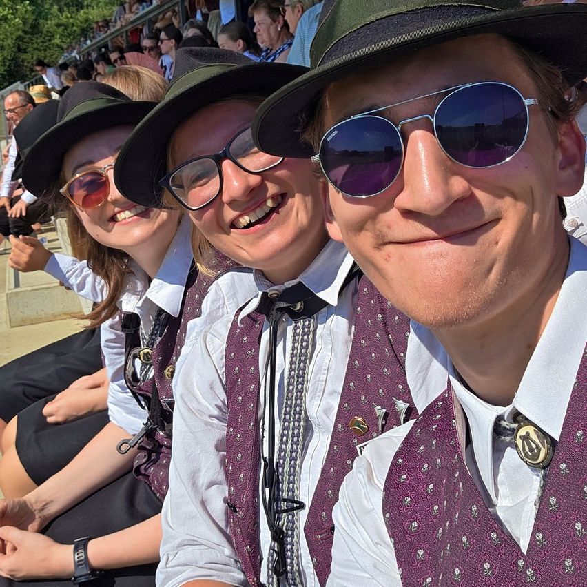 Three people in matching outfits sit together, smiling. They wear hats and sunglasses. The background shows a crowd of people in bleachers.