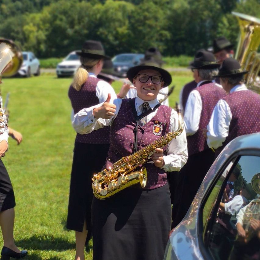 A group of people in hats and vests play instruments on a grassy field. A woman in front smiles and gives a thumbs up while holding a saxophone.