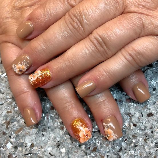 Ein Nahaufnahme der Hände einer Person mit braunen Nägeln, die jeweils mit einem goldenen Blumendesign verziert sind. Die Nägel ruhen auf einer glitzernden Oberfläche.