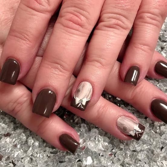 Die Hände einer Person mit braunen Nägeln, einer mit einem weißen Design und einer Schleife, auf einer funkelnden Oberfläche platziert.