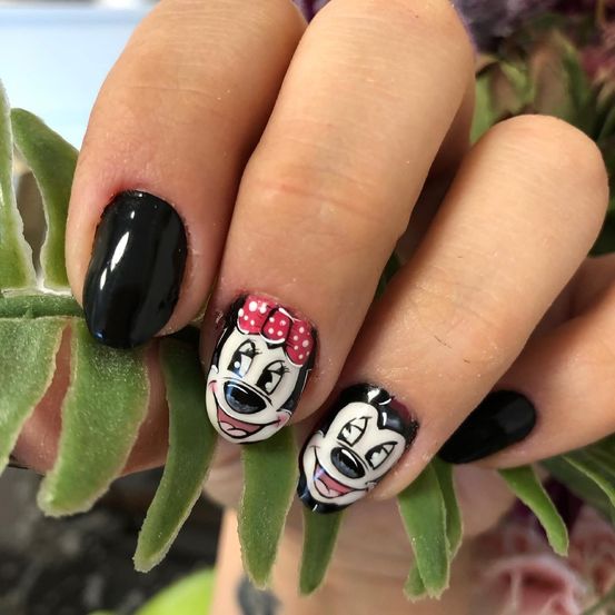 Eine Hand hält einen Fingernagel mit einem Minnie-Maus-Design. Der Fingernagel ist schwarz mit einer rosa Schleife bemalt. Die Nägel werden von grünen Blättern gehalten.