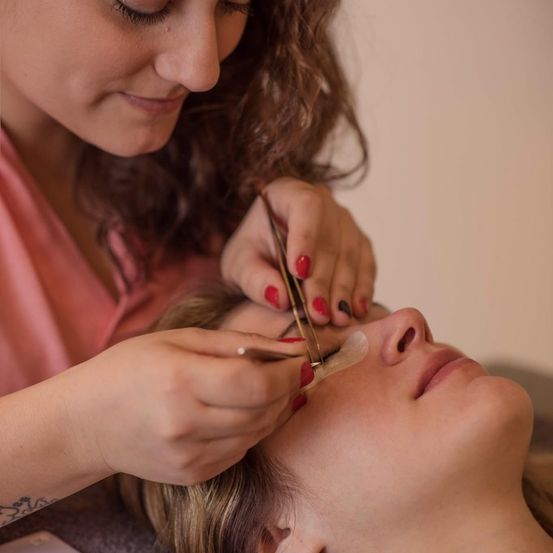 Eine Frau erhält eine Gesichtsbehandlung, mit Pinzetten und Wachspapier auf ihren Augenbrauen. Sie liegt mit geschlossenen Augen auf einer Couch.