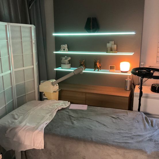 Ein Raum mit einem Bett, beleuchtet von einer Nachttischlampe. Drei Regale sind über einem Holzschrank an der Wand montiert und halten dekorative Gegenstände. Eine weiße Schiebetür befindet sich links.