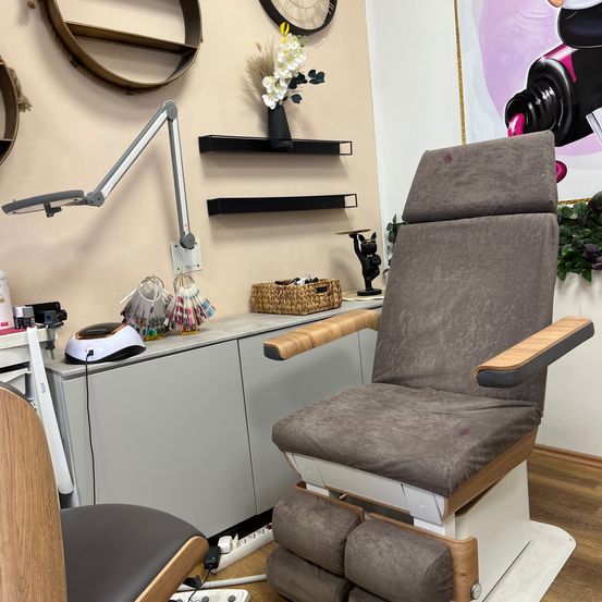 Ein medizinischer Stuhl mit Armlehnen befindet sich in einem Klinikraum. Eine Lampe und eine Blumenvase stehen auf dem Tresen. Eine Steckdose und eine Figur hängen an der Wand. Ein Poster ist an der Wand angebracht.