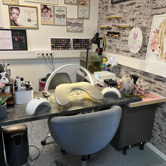 Ein Salon mit einem Schreibtisch, der eine Lampe, einen Maniküresessel und Nagelwerkzeuge hält. Die Wände sind mit gerahmten Zertifikaten und Bildern geschmückt.