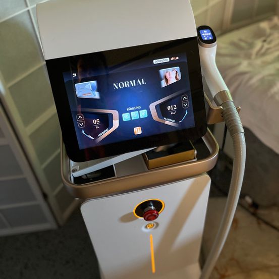 Ein Gerät mit Touchscreen und einem roten Knopf wird gezeigt. Der Bildschirm zeigt 'NORMAL', und ein Frauengesicht ist zu sehen. Es gibt Einstellungen für Kühlung und Frequenz.